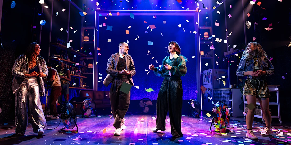 Four people stand and dance on a stage under colorful confetti and dramatic lighting, with props and shelves in the background, suggesting a lively performance scene.