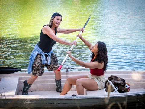 Two women are in a small boat on a body of water, playfully wrestling over a paddle. One stands with a backward cap, while the other is seated. A backpack is beside them.