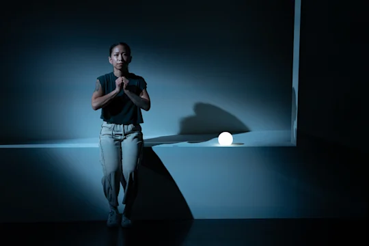 Person standing against a wall in a dimly lit room, hands clasped, next to a glowing white orb on a bench.