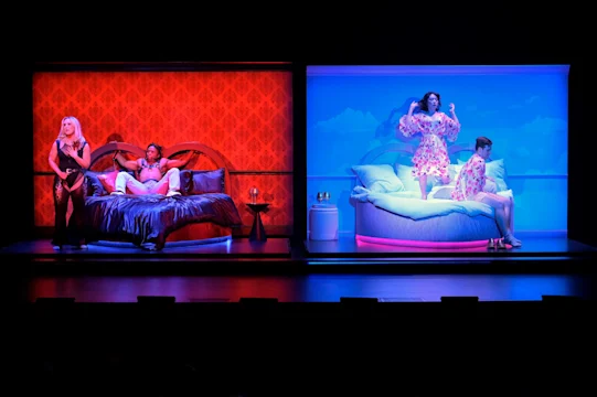 A stage divided into red and blue scenes; two people in bed on the left, and two people in pajamas interacting on the right.