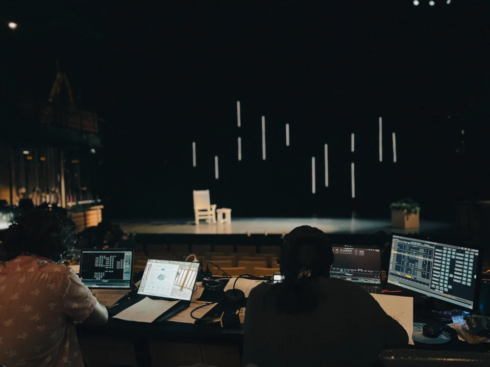 Two individuals manage lighting and sound equipment in a dark theater, facing an empty stage with a single chair and vertical light bars hanging in the background.