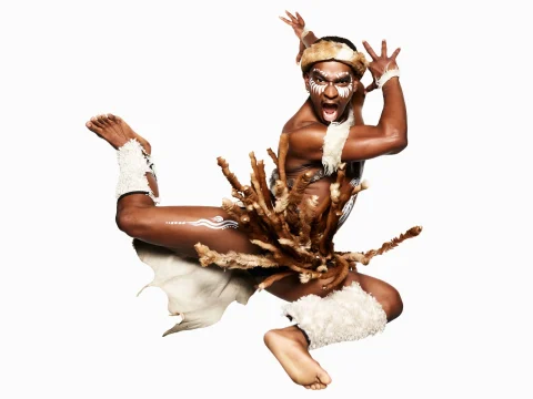 A person in traditional attire with body paint and feather decorations leaps energetically against a plain background.