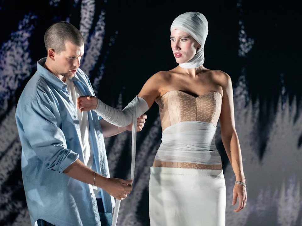A person in a blue shirt wraps bandages around the arm of a woman wearing a nude-colored bodice and a head wrap, both standing against a dark background.