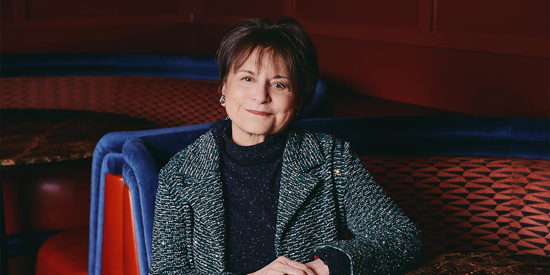A woman with short brown hair and a blue textured jacket sits at a table with red and blue upholstered seating and geometric patterns in the background.