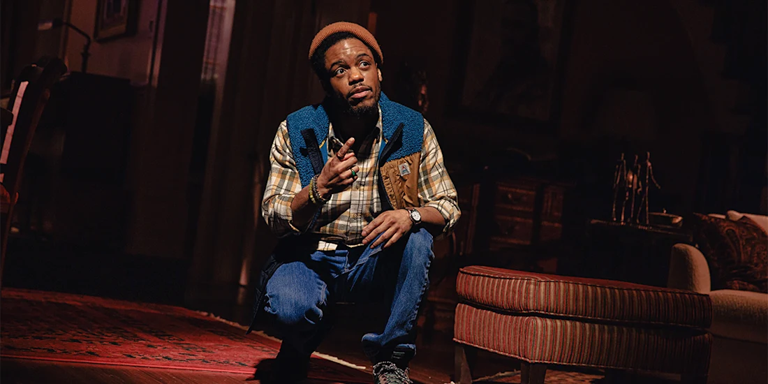 A man wearing a beanie, plaid shirt, and blue vest kneels on the floor indoors, holding a small object and looking upward with a thoughtful expression.