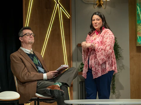 A man in a brown blazer and glasses sits with his eyes closed while a woman in a pink floral top stands and speaks to him on a stage.