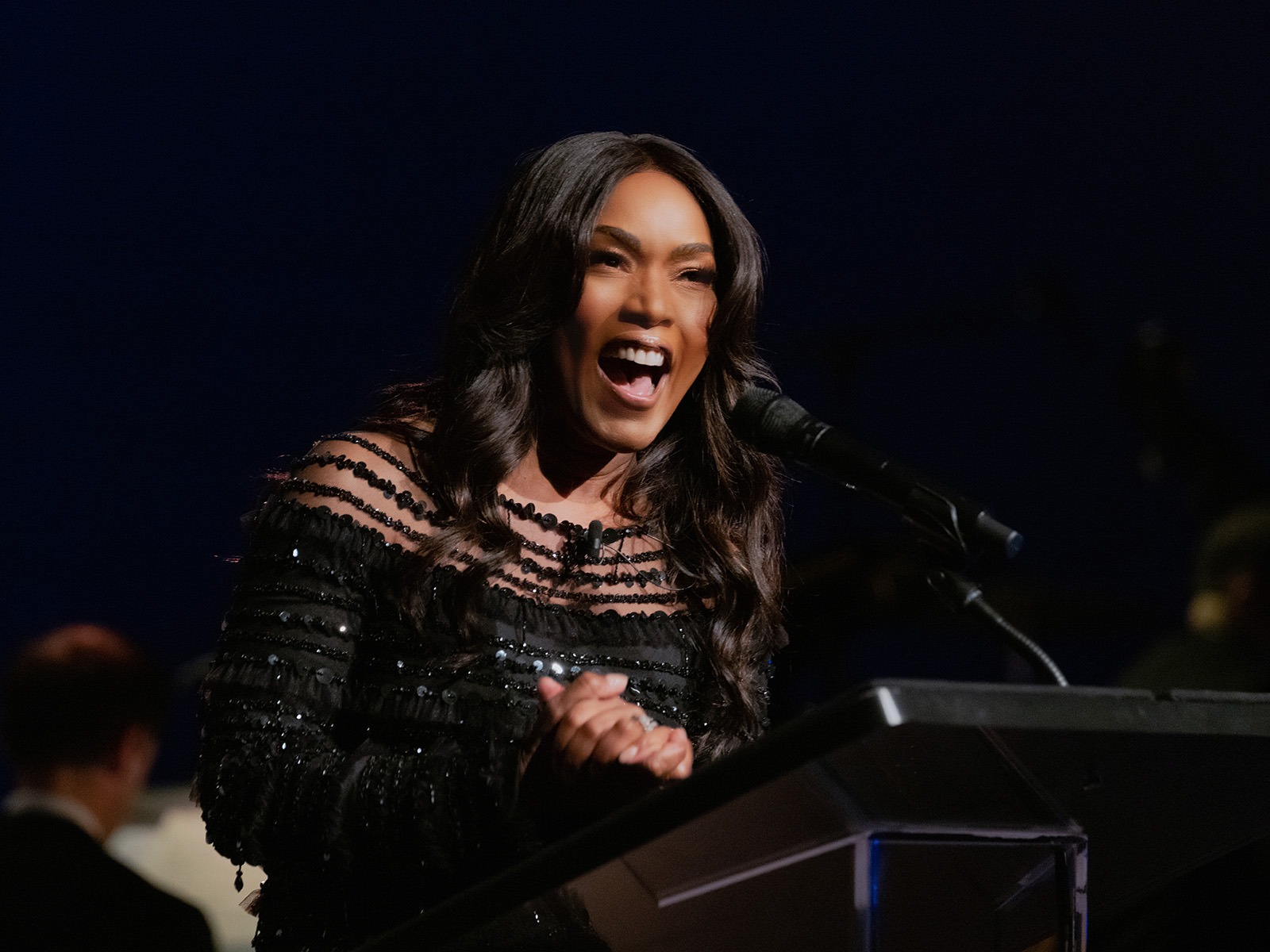 A woman in a black, off-the-shoulder dress speaks animatedly at a podium with a microphone, against a dark background.