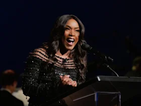 A woman in a black, off-the-shoulder dress speaks animatedly at a podium with a microphone, against a dark background.