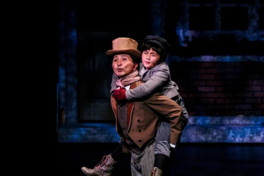 Two actors dressed in historical costumes perform on stage. One carries the other on their back, both wearing hats.