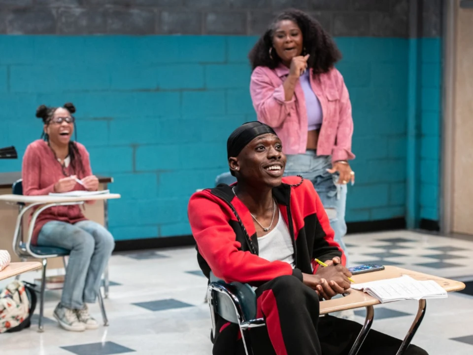 People sitting at desks in a classroom, one person in a red jacket laughing, a person in a pink jacket standing and smiling, and another seated person smiling in the background.