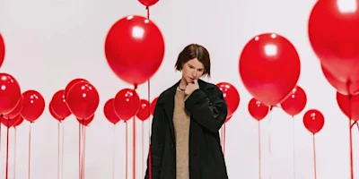 A person with short hair, wearing a black coat and beige sweater, stands among many floating red balloons.