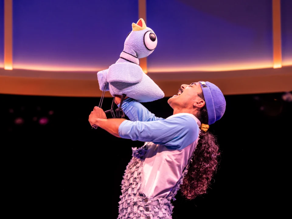 A performer with long hair wearing a blue cap and costume is holding and interacting with a large blue puppet bird in a well-lit stage setting.