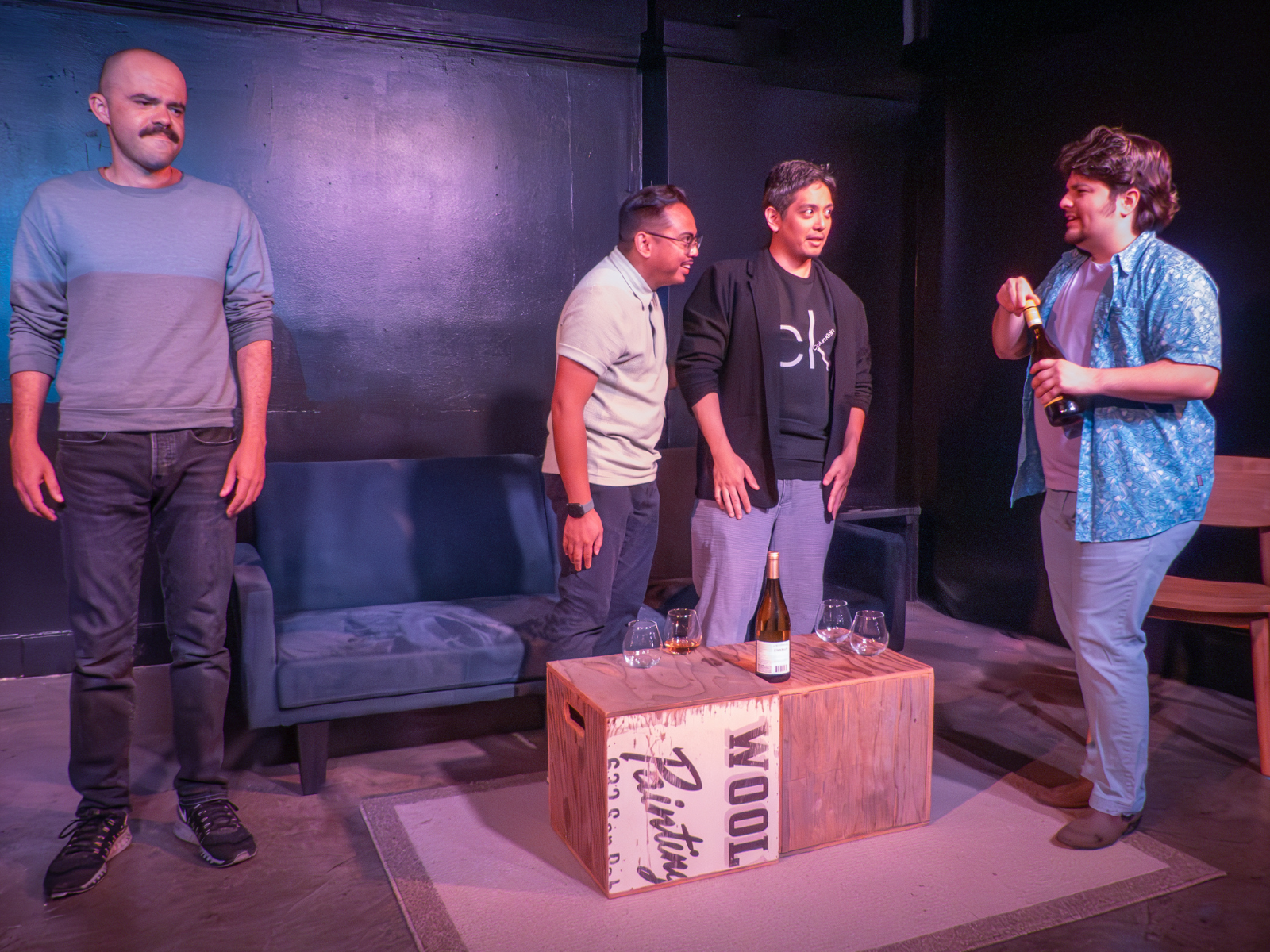 Four individuals are gathered in a room with a sofa, wooden crate, and wine bottle.