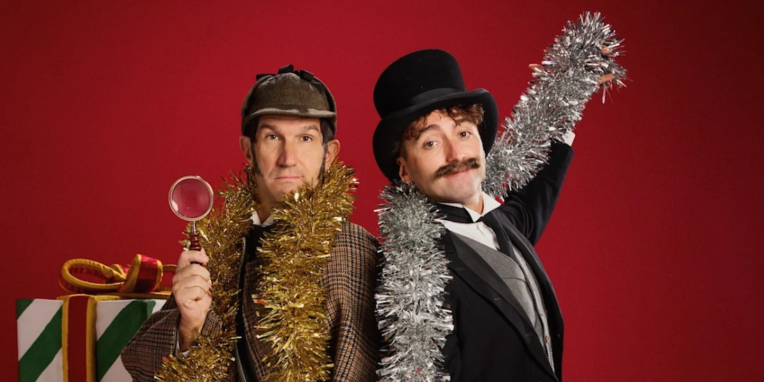 Two men in formal Victorian attire and festive tinsel stand in front of a wrapped gift against a red background; one holds a magnifying glass while the other wears a top hat.