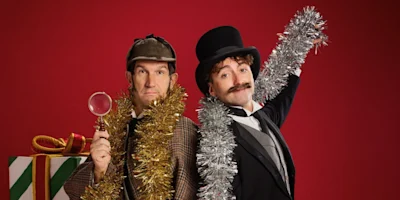 Two men in formal Victorian attire and festive tinsel stand in front of a wrapped gift against a red background; one holds a magnifying glass while the other wears a top hat.