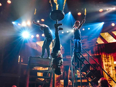 Three performers on stage hold flaming torches; one hangs upside down between two staircases. Bright stage lights and a dramatic set are visible in the background.