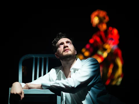 A man in a white shirt leans on a chair, looking upward, with a blurred figure in red illuminated in the background.