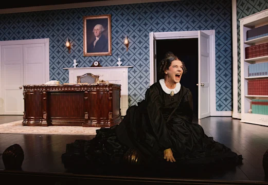 A woman in a black dress kneels on the floor, yelling, in an office set with a large desk, bookshelves, and a portrait of George Washington on the wall.
