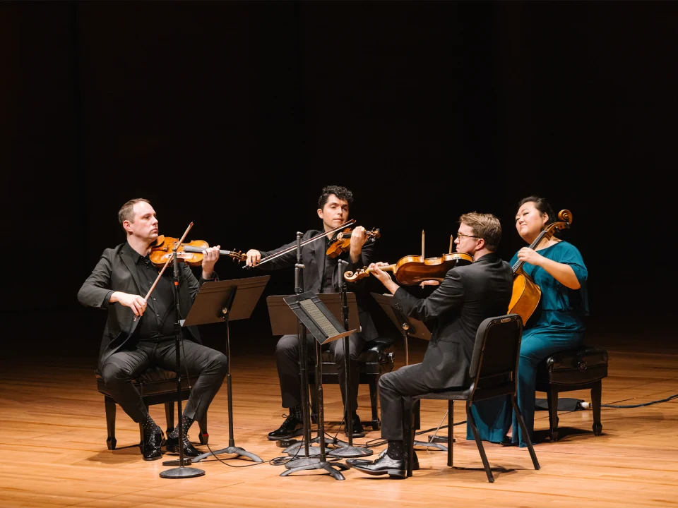 A string quartet performs on stage with three violinists seated in a semicircle and one cellist, all focused on their music.