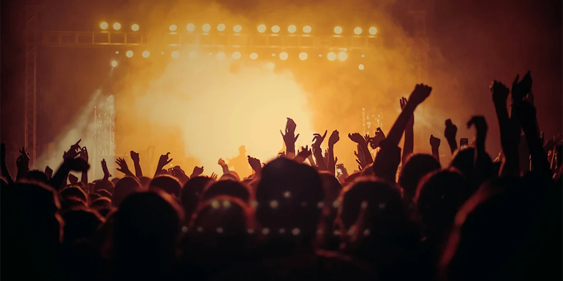 A crowd of people with raised hands faces a brightly lit stage at a concert or music event.