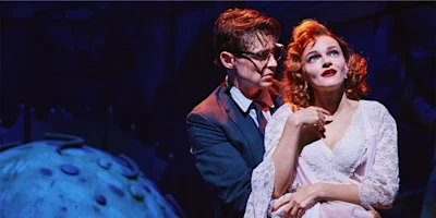 A man in glasses and a suit stands behind a woman in a white dress on stage, under dramatic blue and purple lighting.