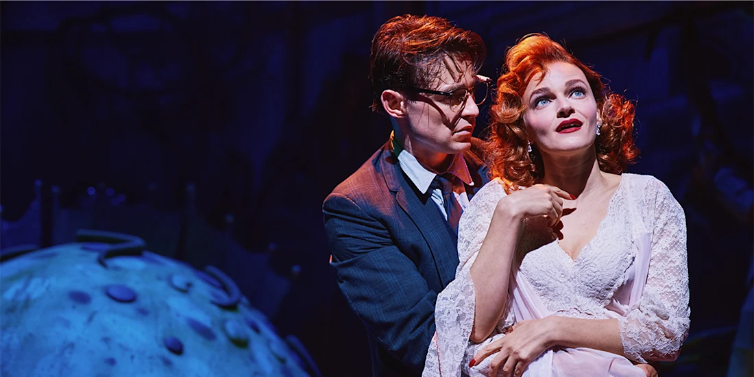 A man in glasses and a suit stands behind a woman in a white dress on stage, under dramatic blue and purple lighting.