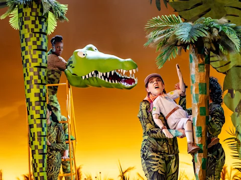 Two performers in green costumes manipulate frog puppets on stage, sitting behind fake rocks, with a large image of the moon projected in the background.