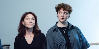 Two people stand side by side against a plain background, one with long brown hair and a black top, the other with short curly hair and a striped shirt, both looking in different directions.