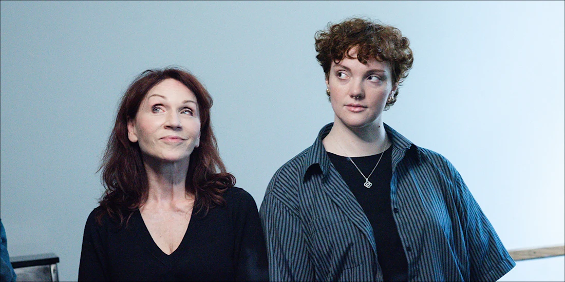 Two people stand side by side against a plain background, one with long brown hair and a black top, the other with short curly hair and a striped shirt, both looking in different directions.
