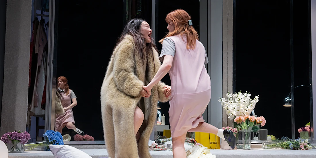 Two women holding hands and laughing in a brightly lit room, one wearing a fur coat and the other in a pink dress, with flowers and a mirror in the background.