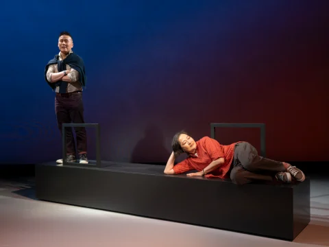 Two individuals on stage: one stands with arms crossed and a jacket over shoulders, the other lies down on the platform, resting their head on a hand. The backdrop displays a gradient red and blue hue.