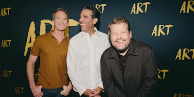 Three men stand smiling and laughing in front of a dark backdrop with the word "ART" repeatedly written in yellow capital letters.