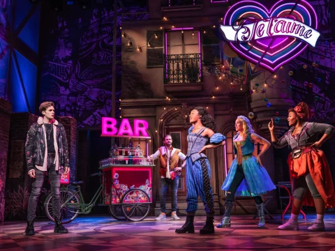 Actors perform a scene on stage in a musical theater production, featuring a bar setup and a neon sign that reads "De Laume" in the background.