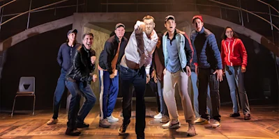A group of seven people on a stage are gathered closely, with one person at the center pointing forward, under dramatic lighting and a modern set design.