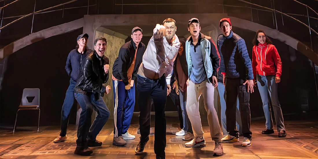 A group of seven people on a stage are gathered closely, with one person at the center pointing forward, under dramatic lighting and a modern set design.