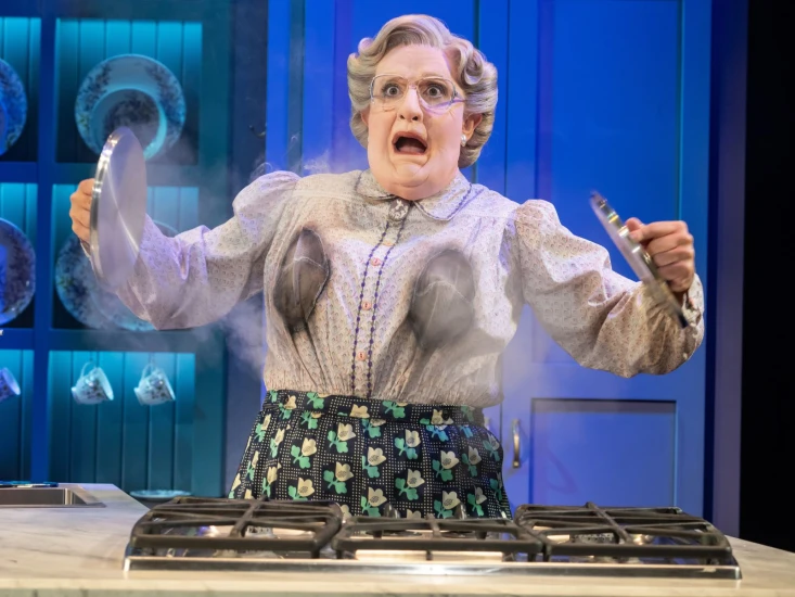 A person dressed in a kitchen outfit with exaggerated makeup and accessories stands in front of a stovetop, looking surprised with smoke rising from their chest area.