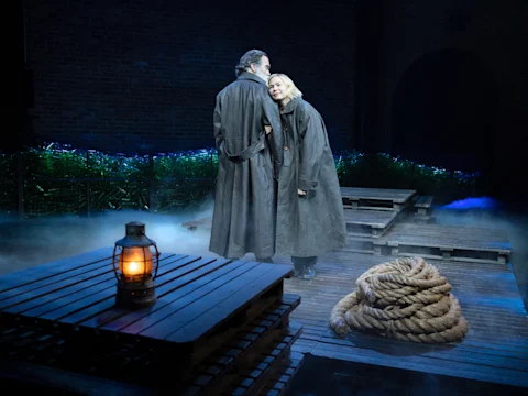 Two actors in long coats stand on a wooden stage set with fog, a coiled rope, and a lit lantern in the foreground. The background features brick walls and greenery.