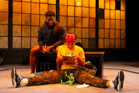 Two people sit on stage; one sits on a bench smiling, while the other sits cross-legged on the floor looking at a tablet, with toys and papers nearby. The background features large windows.