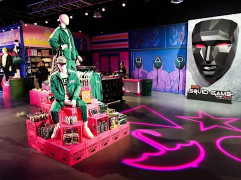 Retail display of Squid Game merchandise, featuring mannequins in green tracksuits, a large mask, and various themed products on pink crates under bright lighting.