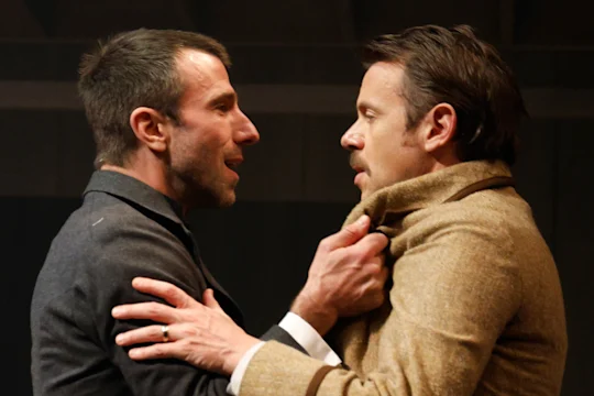 Two men face each other closely, with one gripping the other's coat collar and both appearing to be in an intense conversation.