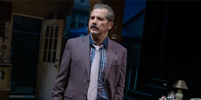 A middle-aged man in a checked suit and tie stands indoors with a serious expression, with a door and stairs visible in the background.