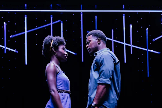 Two people stand face to face under blue lights on a dark stage, gazing intently at each other with serious expressions.