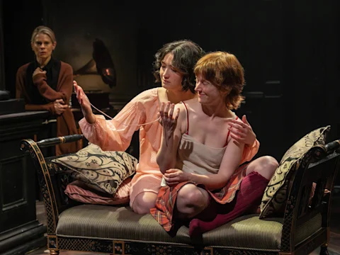Three women are on stage; two sit closely on a couch, one holding a necklace, while a third woman looks on from the background. The scene appears dramatic and intimate.