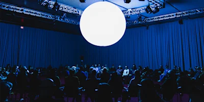 A large, glowing white sphere hangs above a seated audience in a dimly lit, blue-tinted room with curtains covering the walls.