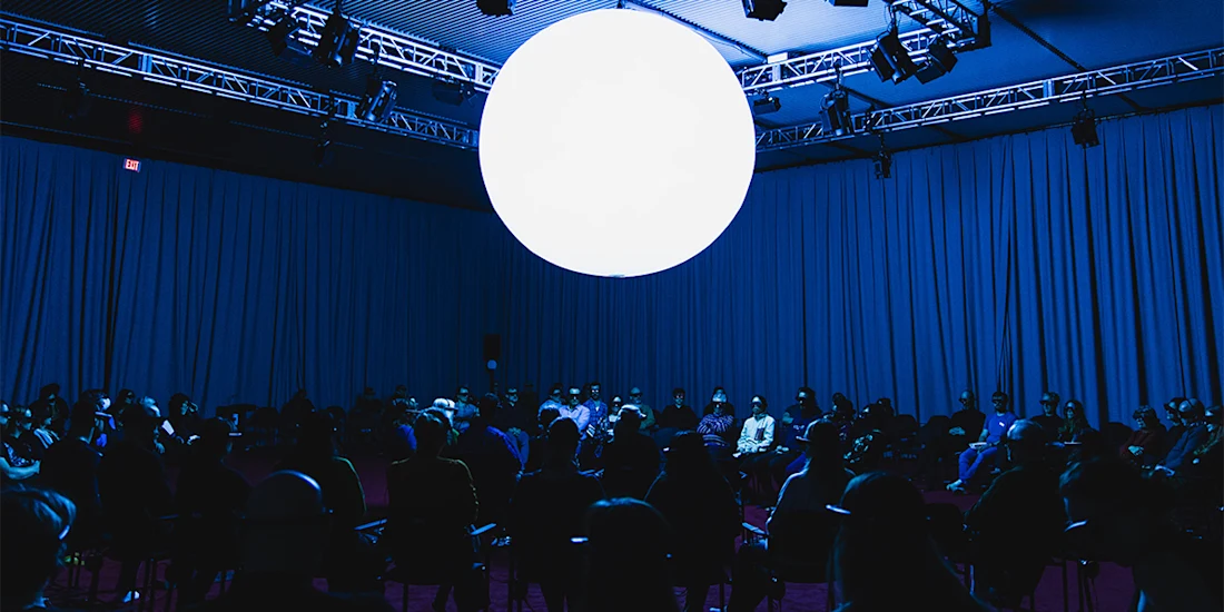 A large, glowing white sphere hangs above a seated audience in a dimly lit, blue-tinted room with curtains covering the walls.
