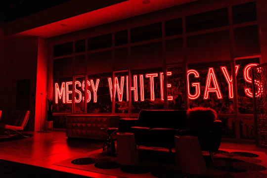 Red neon sign reading "MESSY WHITE GAYS" illuminates a modern living room setup with a couch, chair, and various furniture.