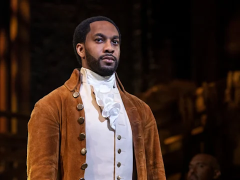 Production photo of Hamilton on Broadway, showing a man dressed in historical 18th-century clothing, including a brown coat and white cravat, stands on stage with a serious expression.