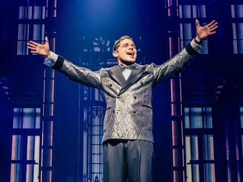 A man in a patterned suit stands center stage with arms outstretched, singing under dramatic lighting and a backdrop of tall windows.