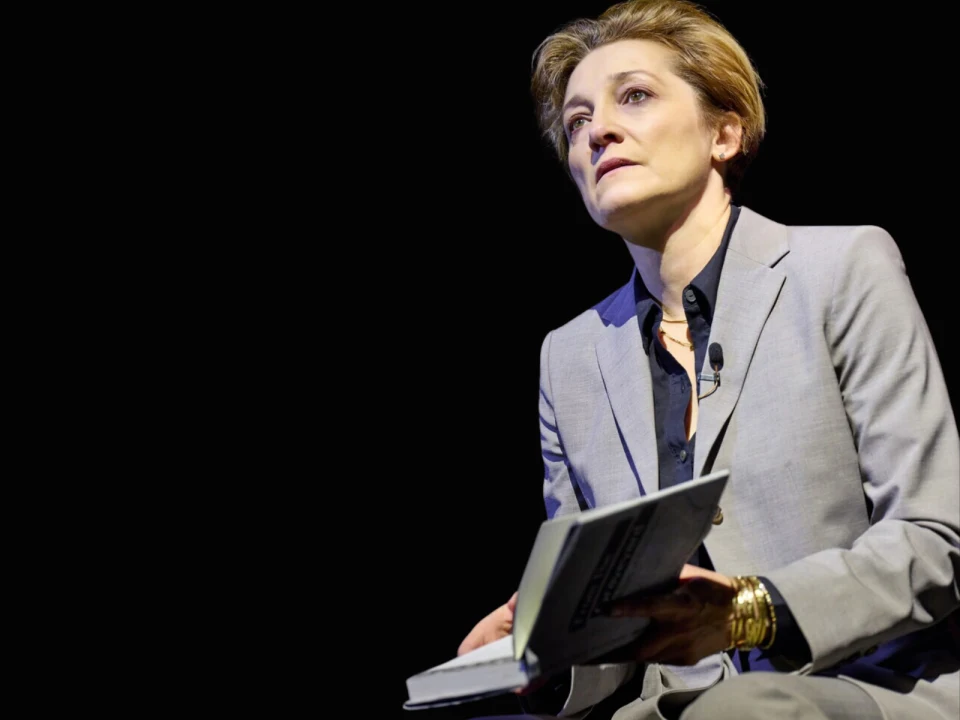 Person in a gray suit sits on stage against a dark background, holding an open book and looking upward.