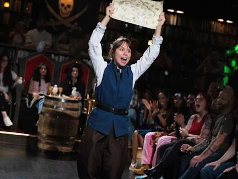 A person in costume holds a map above their head on stage, while an audience seated nearby watches and reacts enthusiastically.